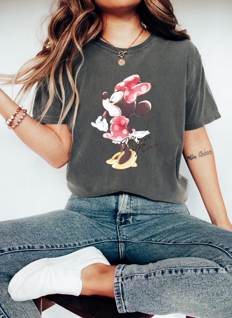 Puede incluir: Una camiseta gris oscuro con un gr&aacute;fico de Minnie Mouse con un vestido rojo de lunares y un lazo rojo. El texto "Minnie Mouse" est&aacute; escrito debajo del gr&aacute;fico.