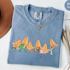 Puede incluir: Camiseta azul vaquero con un gr&aacute;fico de personajes de dibujos animados que llevan conos de tr&aacute;fico naranjas. La camiseta tiene cuello redondo y mangas cortas. Un collar de oro con un colgante est&aacute; drapeado sobre la camiseta.