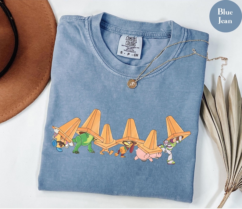 Puede incluir: Camiseta azul vaquero con un gr&aacute;fico de personajes de dibujos animados que llevan conos de tr&aacute;fico naranjas. La camiseta tiene cuello redondo y mangas cortas. Un collar de oro con un colgante est&aacute; drapeado sobre la camiseta.