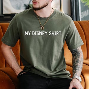 Puede incluir: Camiseta verde oliva con el texto blanco "MY DISNEY SHIRT". La camiseta es de cuello redondo y manga corta. La persona lleva un collar dorado y una pulsera negra.