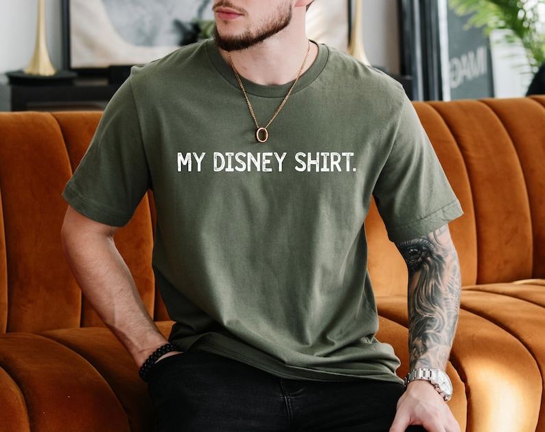 Puede incluir: Camiseta verde oliva con el texto blanco "MY DISNEY SHIRT". La camiseta es de cuello redondo y manga corta. La persona lleva un collar dorado y una pulsera negra.