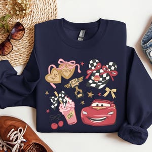 May include: Navy blue sweatshirt featuring cartoon graphics of a red race car, checkered flags, and heart-shaped cookies with the text "KA-CHOW". The design includes pink bows, stars, and a milkshake with the number "95".