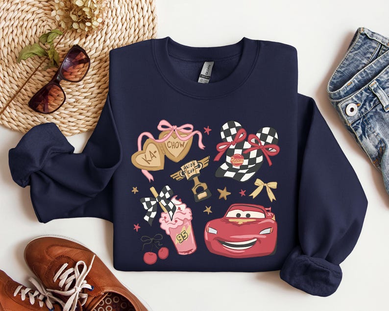 May include: Navy blue sweatshirt featuring cartoon graphics of a red race car, checkered flags, and heart-shaped cookies with the text "KA-CHOW". The design includes pink bows, stars, and a milkshake with the number "95".