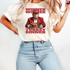 May include: A white t-shirt with a red, white, and blue graphic of a man wearing a red "Make America Great Again" hat holding a turkey. The text "Winner Winner Turkey Dinner" is printed in red on the shirt.