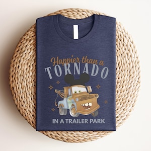 Puede incluir: Una camiseta azul marino con el gr&aacute;fico de una gr&uacute;a marr&oacute;n con orejas de rat&oacute;n negras. El texto "Happier than a TORNADO IN A TRAILER PARK" aparece en fuentes de color naranja y blanco, decorado con peque&ntilde;os iconos de estrellas de cuatro puntas.