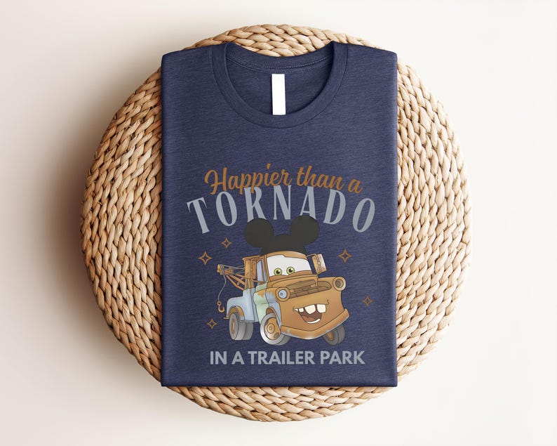 Puede incluir: Una camiseta azul marino con el gr&aacute;fico de una gr&uacute;a marr&oacute;n con orejas de rat&oacute;n negras. El texto "Happier than a TORNADO IN A TRAILER PARK" aparece en fuentes de color naranja y blanco, decorado con peque&ntilde;os iconos de estrellas de cuatro puntas.