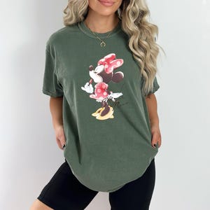 Puede incluir: Una camiseta verde con un gr&aacute;fico de Minnie Mouse con un vestido rojo y un lazo rojo. El texto "Minnie Mouse" est&aacute; impreso debajo del gr&aacute;fico.