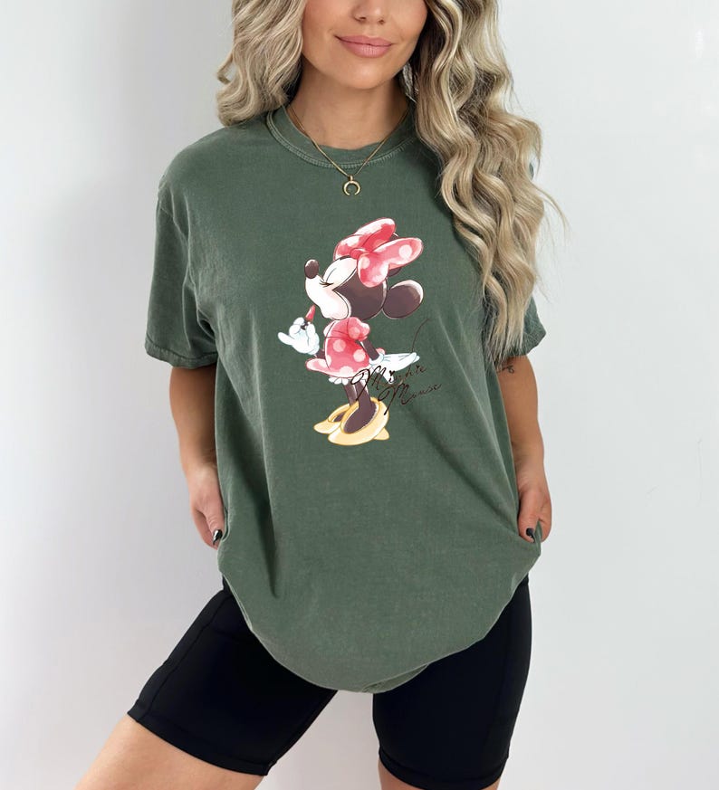 Puede incluir: Una camiseta verde con un gr&aacute;fico de Minnie Mouse con un vestido rojo y un lazo rojo. El texto "Minnie Mouse" est&aacute; impreso debajo del gr&aacute;fico.