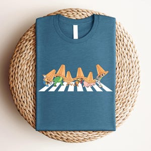 May include: Teal t-shirt featuring a whimsical graphic of cartoon characters wearing orange traffic cones, crossing a white-striped crosswalk. The design is a playful parody. The shirt is folded on a woven, circular surface.