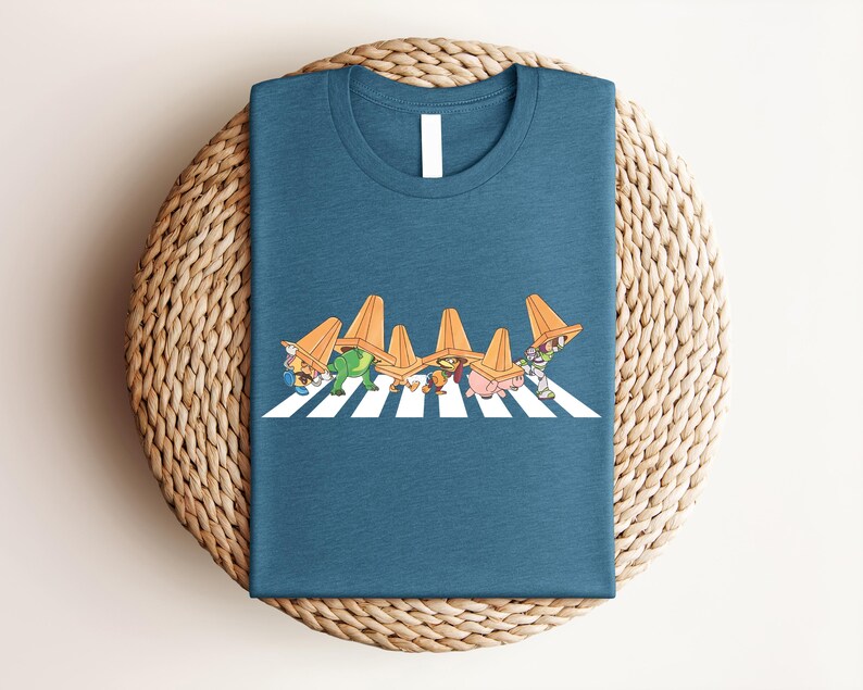 May include: Teal t-shirt featuring a whimsical graphic of cartoon characters wearing orange traffic cones, crossing a white-striped crosswalk. The design is a playful parody. The shirt is folded on a woven, circular surface.