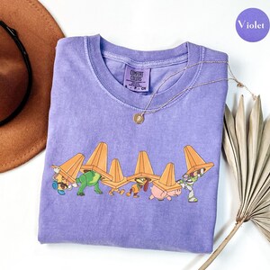 Puede incluir: Una camiseta violeta de cuello redondo Comfort Colors con un gr&aacute;fico de personajes de dibujos animados que llevan conos de tr&aacute;fico naranjas. Un collar de oro con un colgante est&aacute; en la camisa. Un sombrero marr&oacute;n y una planta seca est&aacute;n en el fondo. La palabra "Violet" est&aacute; en un c&iacute;rculo morado.