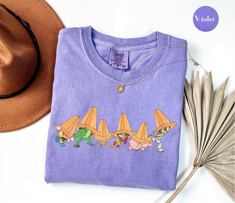Puede incluir: Una camiseta violeta de cuello redondo Comfort Colors con un gr&aacute;fico de personajes de dibujos animados que llevan conos de tr&aacute;fico naranjas. Un collar de oro con un colgante est&aacute; en la camisa. Un sombrero marr&oacute;n y una planta seca est&aacute;n en el fondo. La palabra "Violet" est&aacute; en un c&iacute;rculo morado.