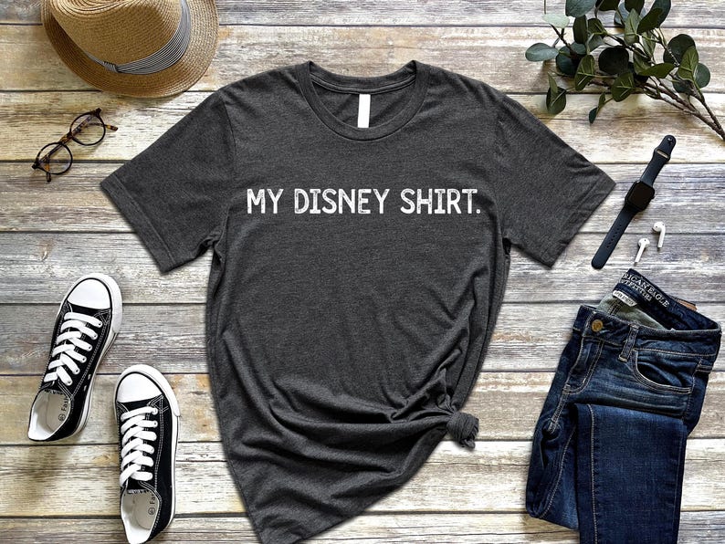 Puede incluir: Camiseta gris oscuro con el texto "MY DISNEY SHIRT". Zapatillas deportivas blancas y negras, vaqueros azules, un sombrero de paja y gafas est&aacute;n colocados sobre una superficie de madera. Tambi&eacute;n se ve un reloj negro y auriculares blancos.