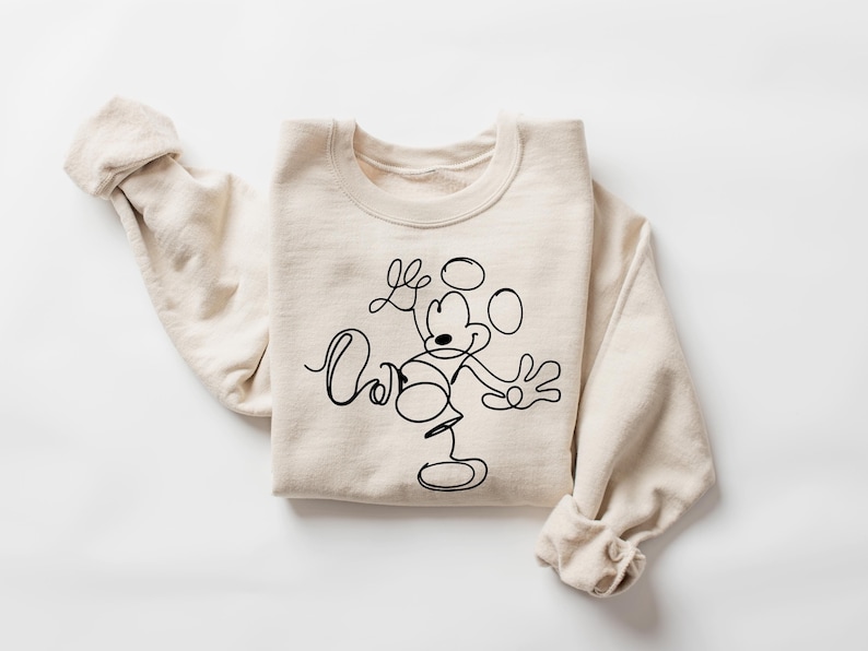 Puede incluir: Una sudadera beige con un dibujo de l&iacute;nea negra de Mickey Mouse haciendo malabares. El dibujo es de estilo minimalista.