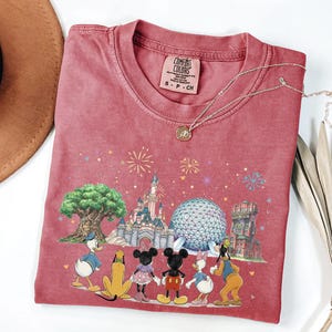 May include: A red t-shirt featuring a colorful graphic of Disney characters and landmarks. The design includes Donald Duck, Mickey and Minnie Mouse, Daisy Duck, and Goofy. The shirt has a crew neck and short sleeves. A gold necklace is draped over the shirt.