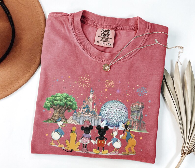 May include: A red t-shirt featuring a colorful graphic of Disney characters and landmarks. The design includes Donald Duck, Mickey and Minnie Mouse, Daisy Duck, and Goofy. The shirt has a crew neck and short sleeves. A gold necklace is draped over the shirt.