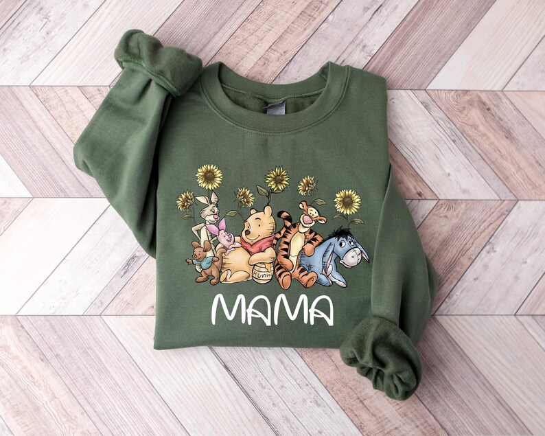 Puede incluir: Sudadera verde con un gr&aacute;fico de Winnie de Pooh y sus amigos, incluyendo Piglet, Tigger, Eeyore y Rabbit. El gr&aacute;fico incluye girasoles y la palabra "MAMA" impresa debajo de los personajes.