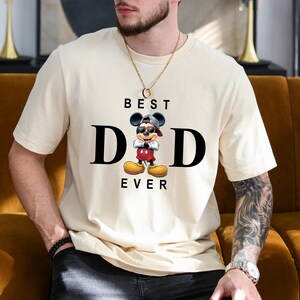 Op de afbeelding: Een wit T-shirt met een zwart-witte afbeelding van Mickey Mouse die een baseballpet achterstevoren draagt en een zonnebril. De tekst "BEST DAD EVER" is onder de afbeelding gedrukt.