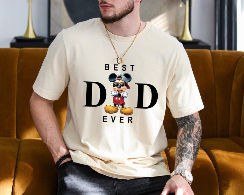 Op de afbeelding: Een wit T-shirt met een zwart-witte afbeelding van Mickey Mouse die een baseballpet achterstevoren draagt en een zonnebril. De tekst "BEST DAD EVER" is onder de afbeelding gedrukt.