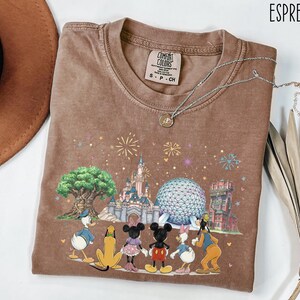 May include: Espresso-colored t-shirt featuring a colorful graphic of Disney characters and landmarks. The design includes Donald Duck, Pluto, Mickey Mouse, Minnie Mouse, Daisy Duck, and Goofy. The shirt has a crew neck and short sleeves.