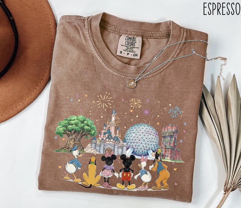 May include: Espresso-colored t-shirt featuring a colorful graphic of Disney characters and landmarks. The design includes Donald Duck, Pluto, Mickey Mouse, Minnie Mouse, Daisy Duck, and Goofy. The shirt has a crew neck and short sleeves.