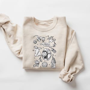K&ouml;nnte beinhalten: Ein cremefarbenes Sweatshirt mit einem schwarz-wei&szlig;en Grafikdesign. Das Design zeigt verschiedene Charaktere und Elemente aus einem beliebten Animationsfilm, wie einen Hexenhut, einen Geist und ein Charakterportr&auml;t. Das Sweatshirt hat lange &Auml;rmel.
