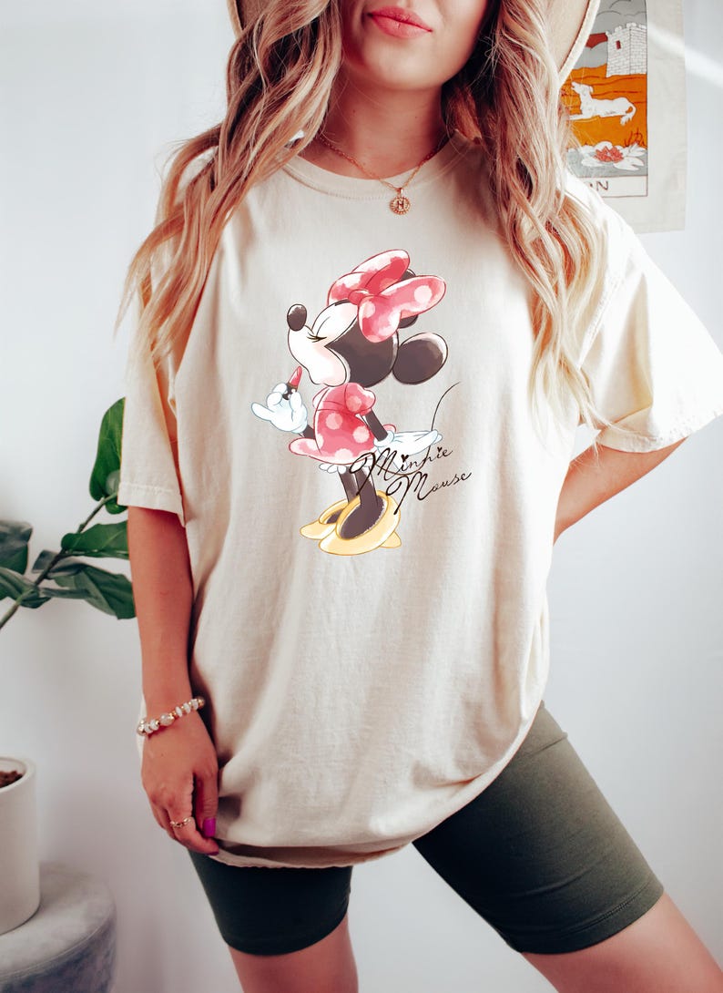 Puede incluir: Una camiseta de color crema con un gr&aacute;fico de Minnie Mouse con un vestido rojo con lunares blancos y un lazo rojo. El texto "Minnie Mouse" est&aacute; escrito en cursiva negra debajo del gr&aacute;fico.