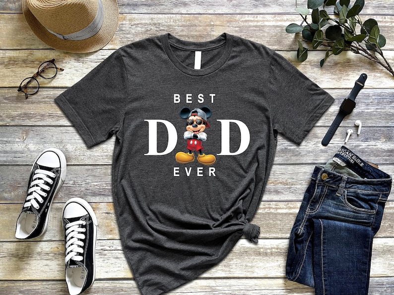 Op de afbeelding: Een donkergrijs T-shirt met een witte afbeelding van Mickey Mouse met een zonnebril en een petje achterstevoren. De tekst "BEST DAD EVER" is in wit onder de afbeelding gedrukt.