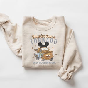 Puede incluir: Una sudadera beige de cuello redondo con el gr&aacute;fico de una gr&uacute;a marr&oacute;n y una silueta de orejas de rat&oacute;n negras encima. El texto "Happier than a TORNADO IN A TRAILER PARK" aparece en letras marrones y azules, decorado con peque&ntilde;as estrellas.