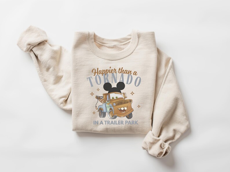 Puede incluir: Una sudadera beige de cuello redondo con el gr&aacute;fico de una gr&uacute;a marr&oacute;n y una silueta de orejas de rat&oacute;n negras encima. El texto "Happier than a TORNADO IN A TRAILER PARK" aparece en letras marrones y azules, decorado con peque&ntilde;as estrellas.