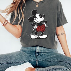 May include: A gray t-shirt with a vintage-style Mickey Mouse graphic. The shirt is worn by a person wearing blue jeans and white sneakers.