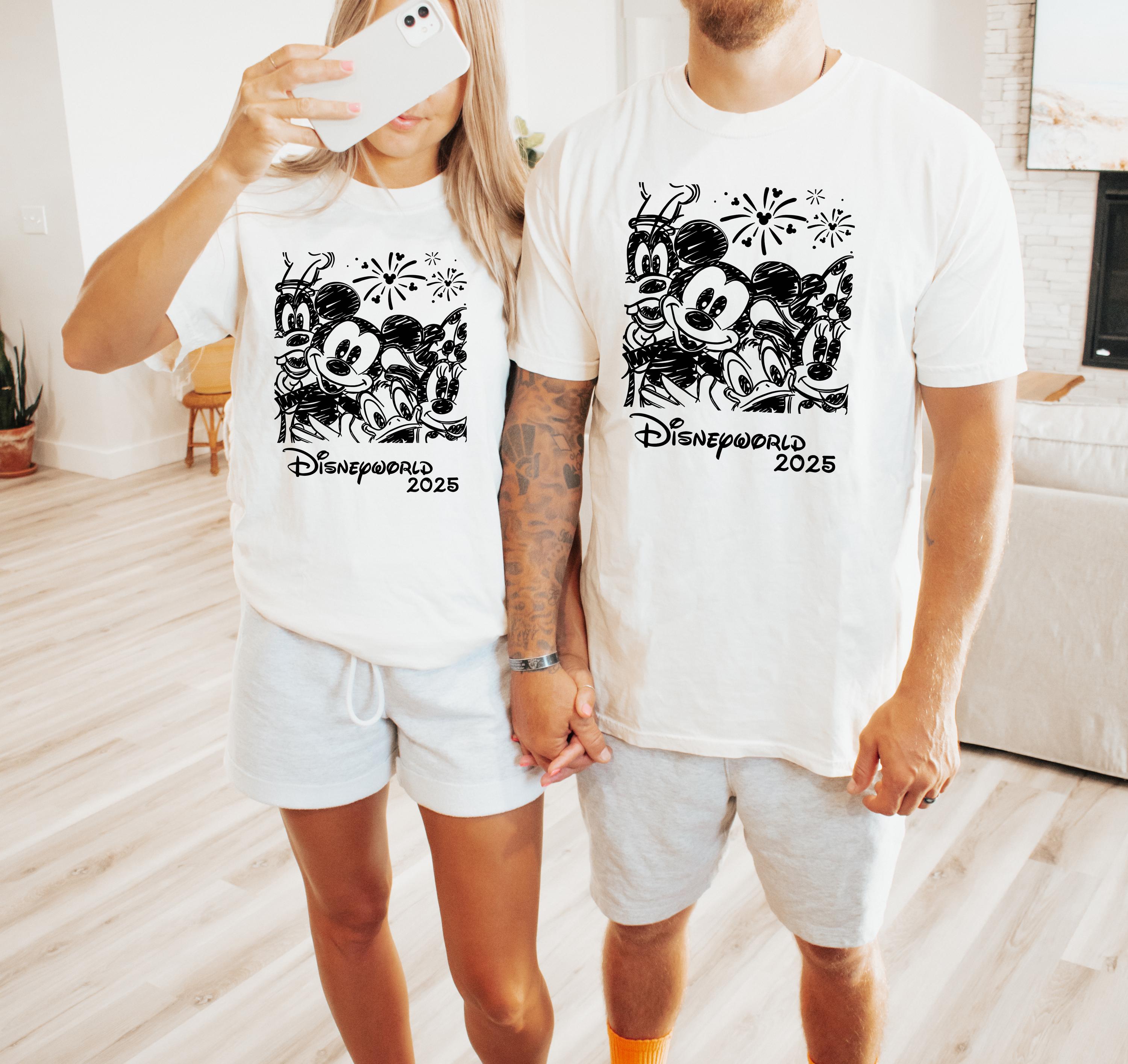 K&ouml;nnte beinhalten: Ein Paar tr&auml;gt wei&szlig;e T-Shirts mit einer Schwarzwei&szlig;illustration von Mickey Mouse, Donald Duck und Goofy mit Feuerwerk. Der Text "Disneyworld 2025" ist auf den Shirts gedruckt.