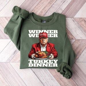May include: Green sweatshirt with a white graphic of a person wearing a red hat with the text "Make America Great Again" on it. The person is holding a plate with a roasted turkey on it. The text "Winner Winner Turkey Dinner" is printed on the sweatshirt.