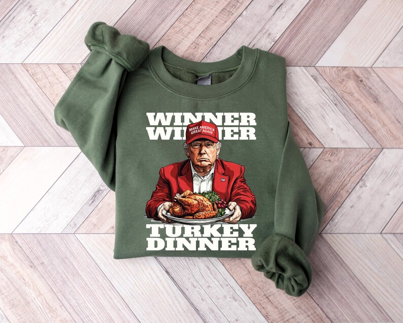 May include: Green sweatshirt with a white graphic of a person wearing a red hat with the text "Make America Great Again" on it. The person is holding a plate with a roasted turkey on it. The text "Winner Winner Turkey Dinner" is printed on the sweatshirt.