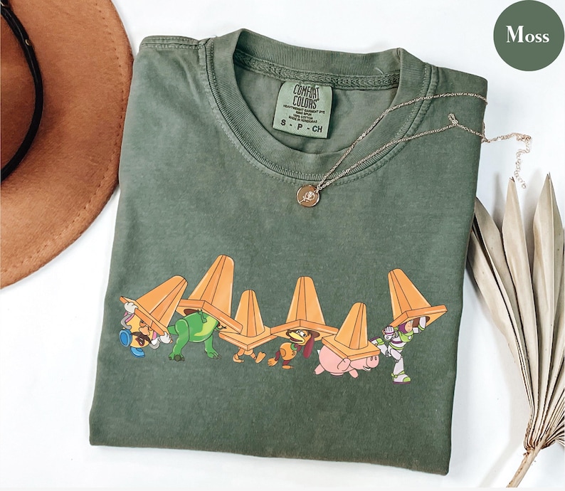 Puede incluir: Camiseta verde musgo con un gr&aacute;fico de personajes de dibujos animados que llevan conos de tr&aacute;fico naranjas. La camiseta tiene cuello redondo y mangas cortas. Un collar dorado con un colgante est&aacute; sobre la camiseta.