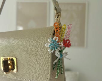 May include: A beige leather handbag with a gold clasp and a beaded flower bouquet charm. The charm features colorful beads in shades of blue, orange, red, and green, attached to a gold-tone clasp.