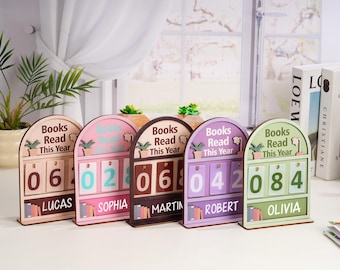 May include: A row of wooden book counters in various colors, each displaying a different number of books read. Each counter has the text "Books Read This Year" and a name at the bottom. The counters are in shades of brown, pink, purple, and green.