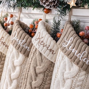 May include: Four knit Christmas stockings in alternating cream and beige colors, each featuring a folded cuff with embroidered names Ribon, Justin, Kevin, and Linda. The stockings have a cable knit pattern and are decorated with pine and berries.