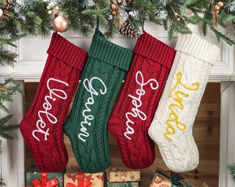 May include: Four cable-knit Christmas stockings in red, green, and white, each with a name embroidered in a contrasting color: Violet, Gavin, Sophia, and Linda. The stockings hang from a decorated mantel.