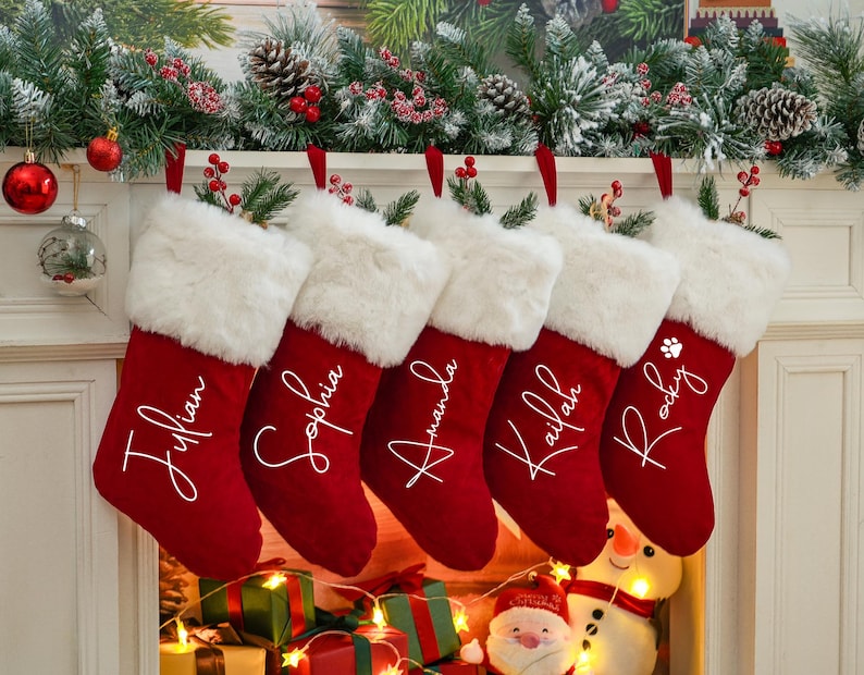 May include: Five red velvet Christmas stockings with white faux fur trim hanging on a fireplace mantel. The stockings are personalized with the names "Julian", "Sophia", "Amanda", "Kailan", and "Rocky".