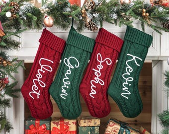 May include: Four personalized Christmas stockings in red and green, each with a name in white script: Violet, Gavin, Sophia, and Kevin. The stockings are hanging above wrapped gifts and festive greenery.