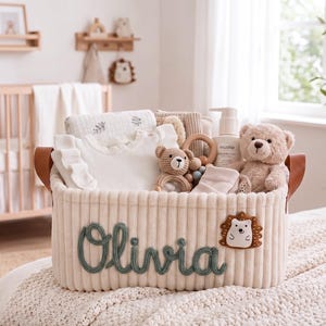 Peut inclure: Un panier de rangement en tissu côtelé crème avec des poignées en similicuir marron. Le prénom Olivia est brodé en écriture verte sur le devant, accompagné d'un petit écusson hérisson. Le panier contient une chemise blanche à volants, un ours en peluche, un hochet en bois et un flacon de lotion pour bébé Mushie.