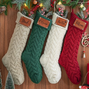 Könnte beinhalten: Vier personalisierte, gestrickte Weihnachtsstrümpfe mit den Namen "Papa", "Olivia", "Bella" und "Mama" auf braunen Lederetiketten. Die Strümpfe sind cremefarben, grün, weiß und rot.