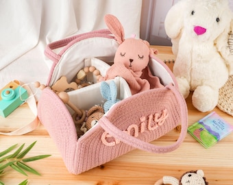 May include: A pink, heart-shaped baby bag with the name "Emily" embroidered on the side, filled with baby toys, including a plush bunny, wooden toys, and a small blue knitted bunny. A large white stuffed bunny is in the background.
