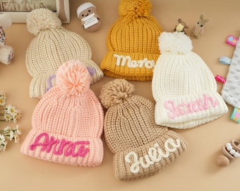 Può includere: Cinque berretti di lana con pon pon e nomi personalizzati. I berretti sono di vari colori, tra cui beige, bianco, rosa, marrone e giallo. I nomi sui berretti sono "Arlo", "Julia", "Sarah", "Mateo" e "Arlo".
