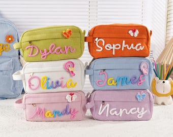 May include: A stack of six corduroy pencil cases in various colors, each with a name embroidered on the front. The colors include green, orange, white, blue, and pink. Decorative elements include butterflies and a lollipop.