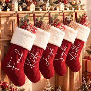 Può includere: Cinque calze natalizie in velluto rosso con risvolti in pelliccia sintetica bianca, personalizzate con testo in corsivo bianco: Frank, Kirsten, Joseph, Sophie e Arthur. Ogni calza è riempita con scatole regalo, pino e bacche rosse.