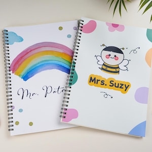 Może przedstawiać: Dwa spiralne notatniki. Jeden przedstawia akwarelową tęczę i imię "Mr. Patrick". Drugi ma kreskówkową pszczołę z tekstem "Mrs. Suzy". Widoczne są kolorowe akcenty i ołówek.
