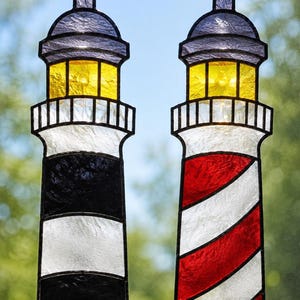 May include: Two stained glass lighthouse suncatchers hanging from metal chains. The left lighthouse features horizontal black and white stripes, while the right lighthouse has diagonal red and white stripes. Both have yellow lanterns and purple roofs.
