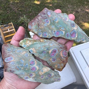 May include: Three irregular, flat slices of ruby in fuchsite mineral stone. Each piece features a mottled sage green base with embedded circular inclusions of vibrant magenta-pink ruby crystals surrounded by rings of light blue kyanite.